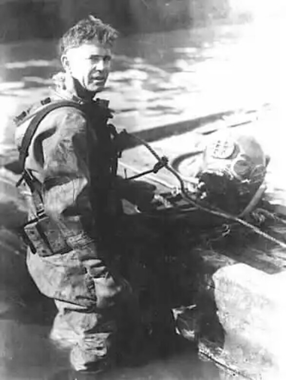 A man in dive gear stands in water up to his knees.