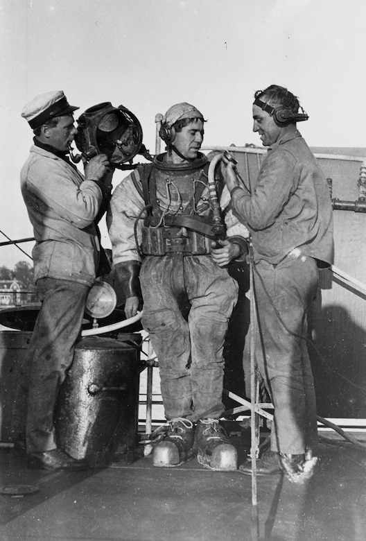 Two men help a third man to get into early 1900s diving gear.