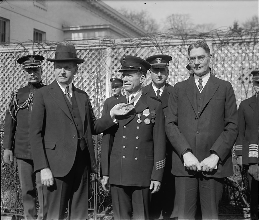 Three men stand beside each other. One holds a medal on the other man’s neck.