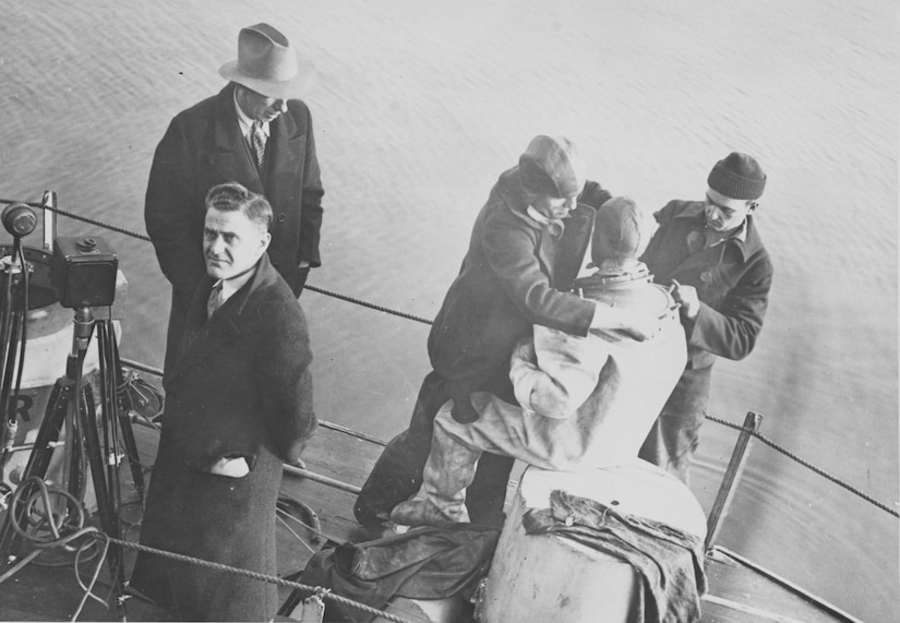 Two men dress a third man in diving gear. Two other men stand near a camera.