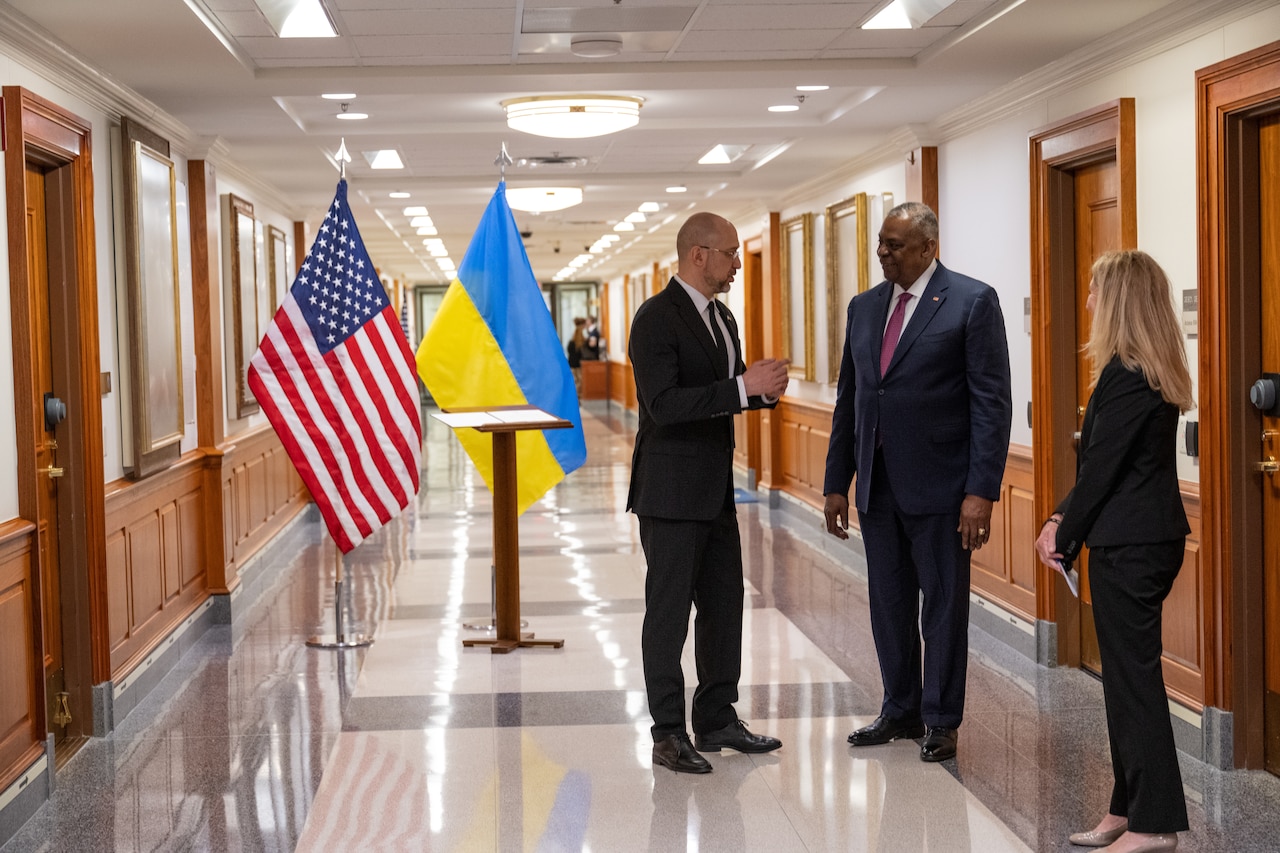 Two men in business attire speak informally in an office building hallway.