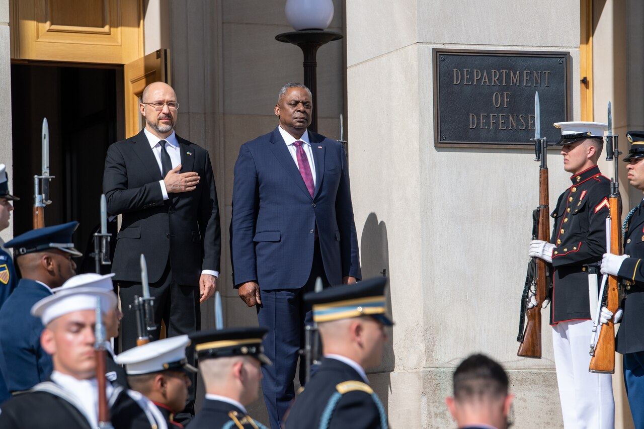 Two men in business attire stand among others in military dress. The sign on the building indicates that they are at the Department of Defense.