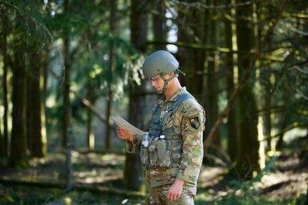 A Soldier standing in a forest looking at a map wearing helmet and eye protection.