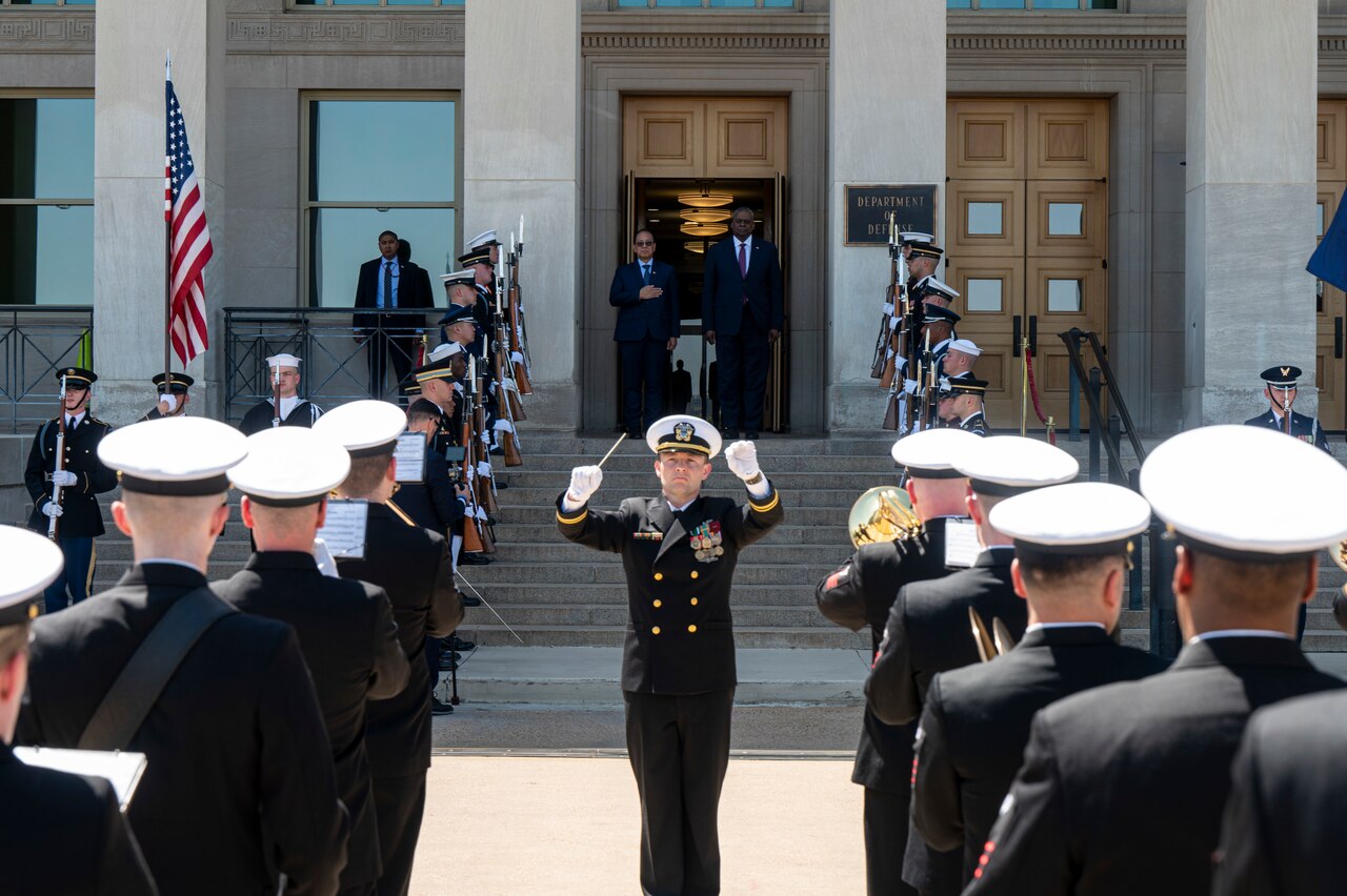 Two men stand at the top of stairs. A military band  is in the foreground.