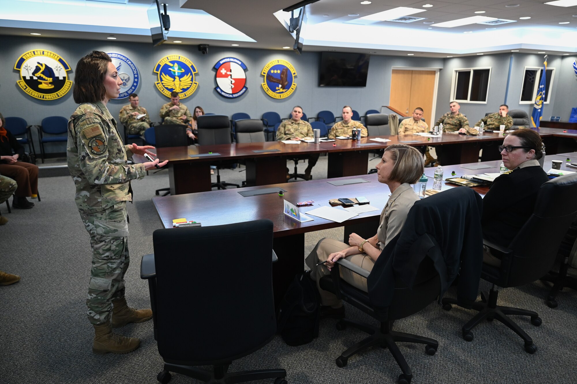 Maj. stands at the front of the room as other military members sit around a horse shoe table.