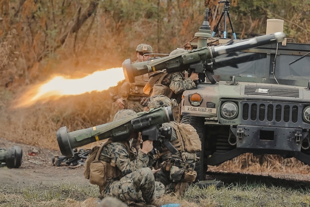 Fire shoots from the back of a shoulder launcher as one of several soldiers behind a vehicle launches a missile in the woods.