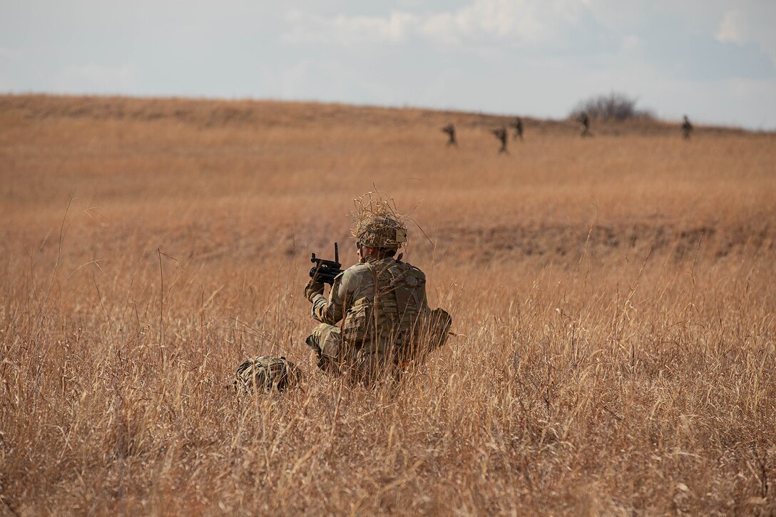 A soldier holds a weapon while squatting in a field while other soldiers hold their weapons in the background.