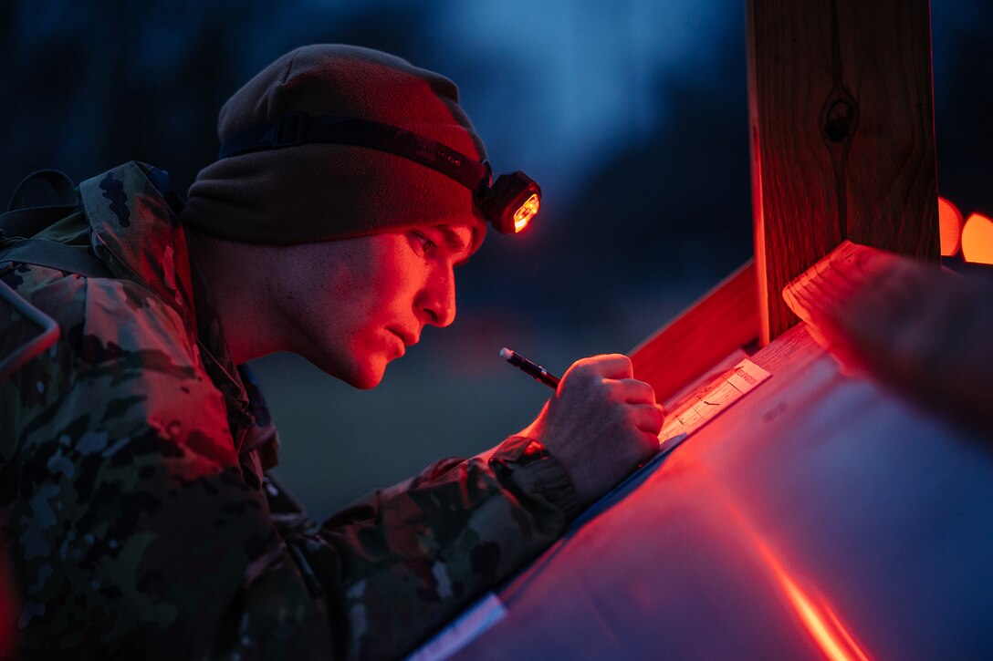Close-up photo of a soldier writing at night, illuminated by a red light on the soldier’s headband.