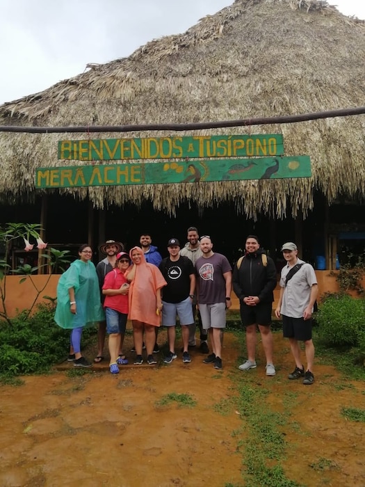 Airmen in civilian clothes and ponchos stand under a straw roof and sign that reads "Bienvenidos a Tusipono"
