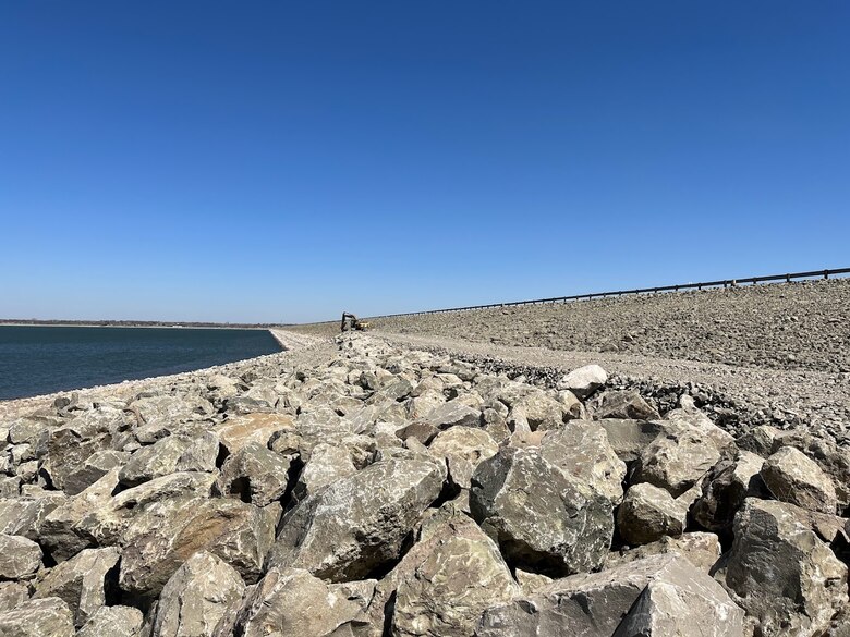 A group of five individuals stand on rock being placed on the face of the Harlan County Dam and inspect the work being done. There is a blue sky above and water in the left background.