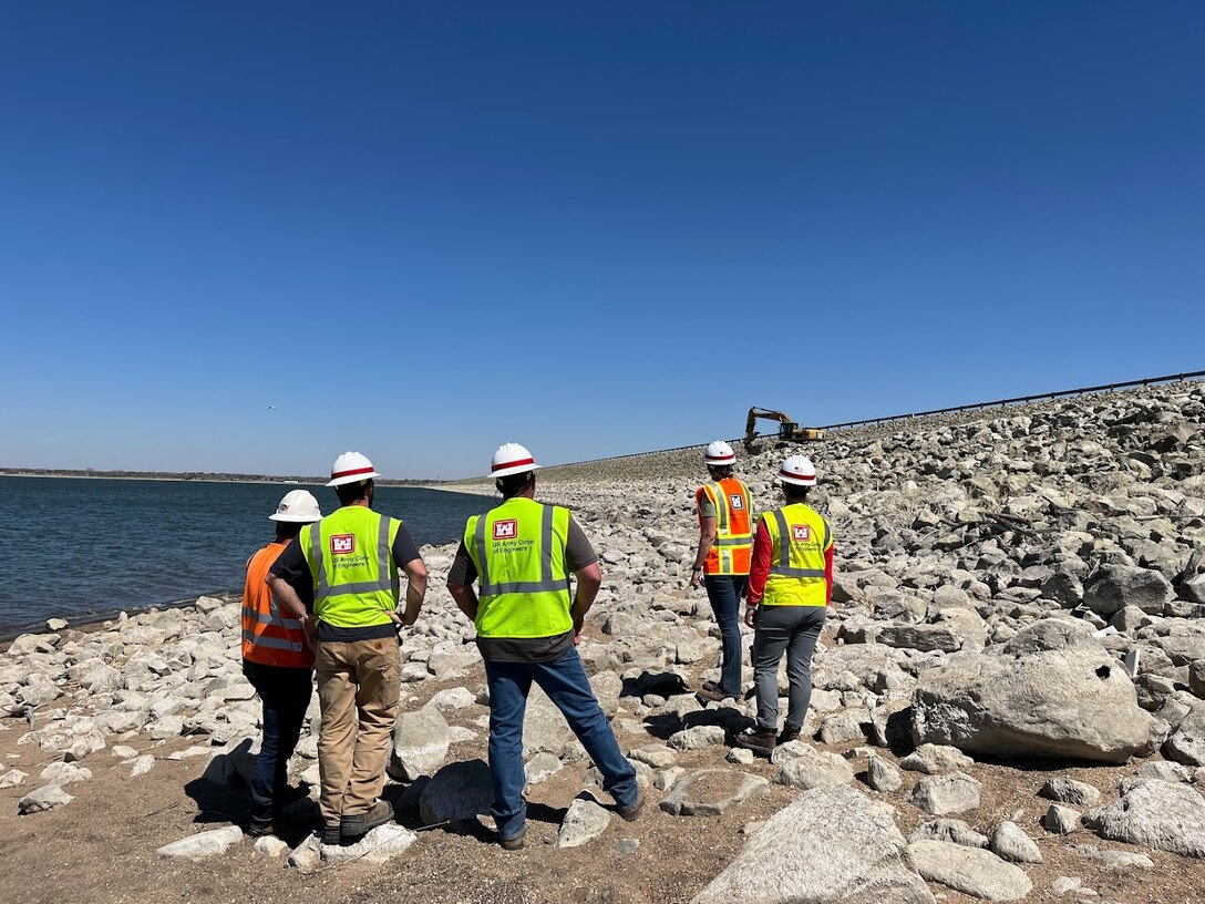 A group of five individuals stand on rock being placed on the face of the Harlan County Dam and inspect the work being done. There is a blue sky above and water in the left background.