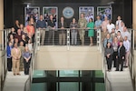 A group of people stand on a split staircase in a federal government building. Military photos are behind them.