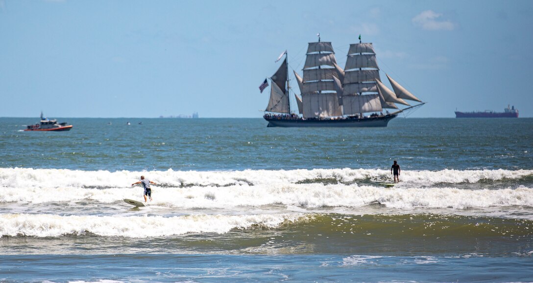 Surfers ride waves as Elissa, a three-masted barque and one of three ships of her kind still active, transits the Gulf of Mexico off the coast of Galveston Beach during Tall Ships Challenge Galveston 2023.
