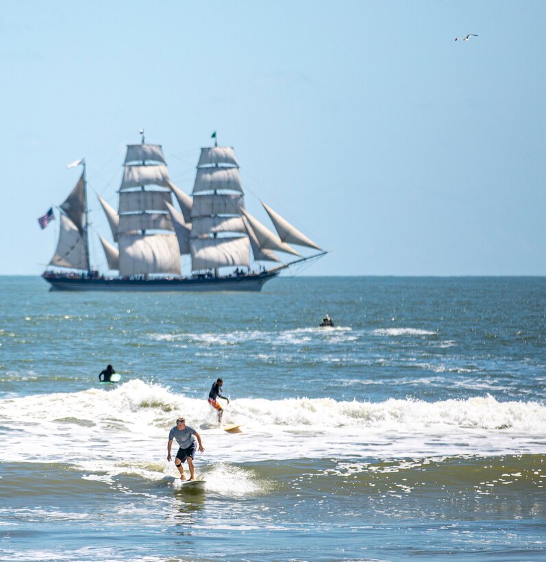 Surfers ride waves as Elissa, a three-masted barque and one of three ships of her kind still active, transits the Gulf of Mexico off the coast of Galveston Beach during Tall Ships Challenge Galveston 2023.