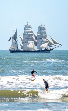 Surfers ride waves as Elissa, a three-masted barque and one of three ships of her kind still active, transits the Gulf of Mexico off the coast of Galveston Beach during Tall Ships Challenge Galveston 2023.