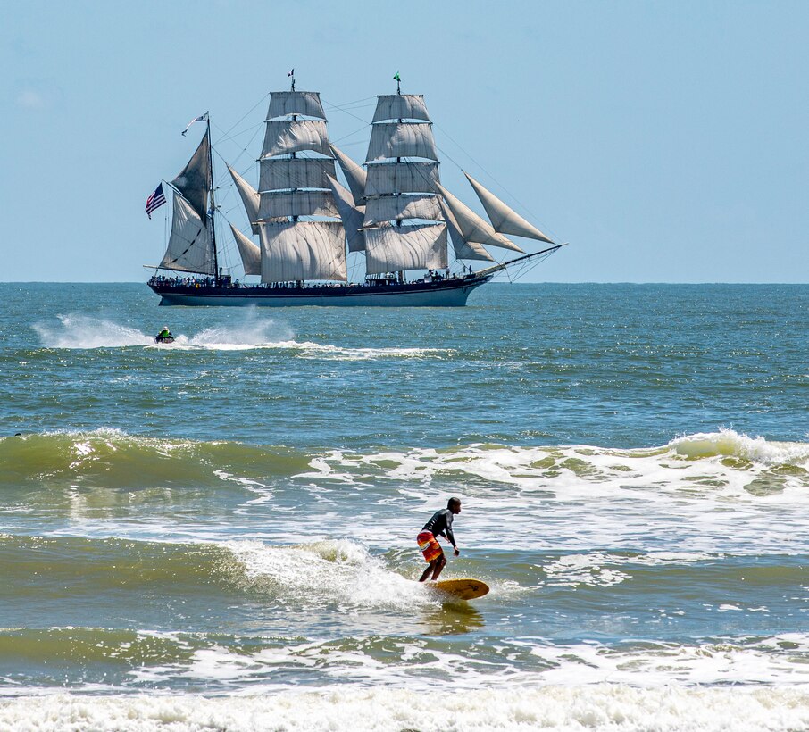 A surfer rides a wave as Elissa, a three-masted barque and one of three ships of her kind still active, transits the Gulf of Mexico off the coast of Galveston Beach during Tall Ships Challenge Galveston 2023
