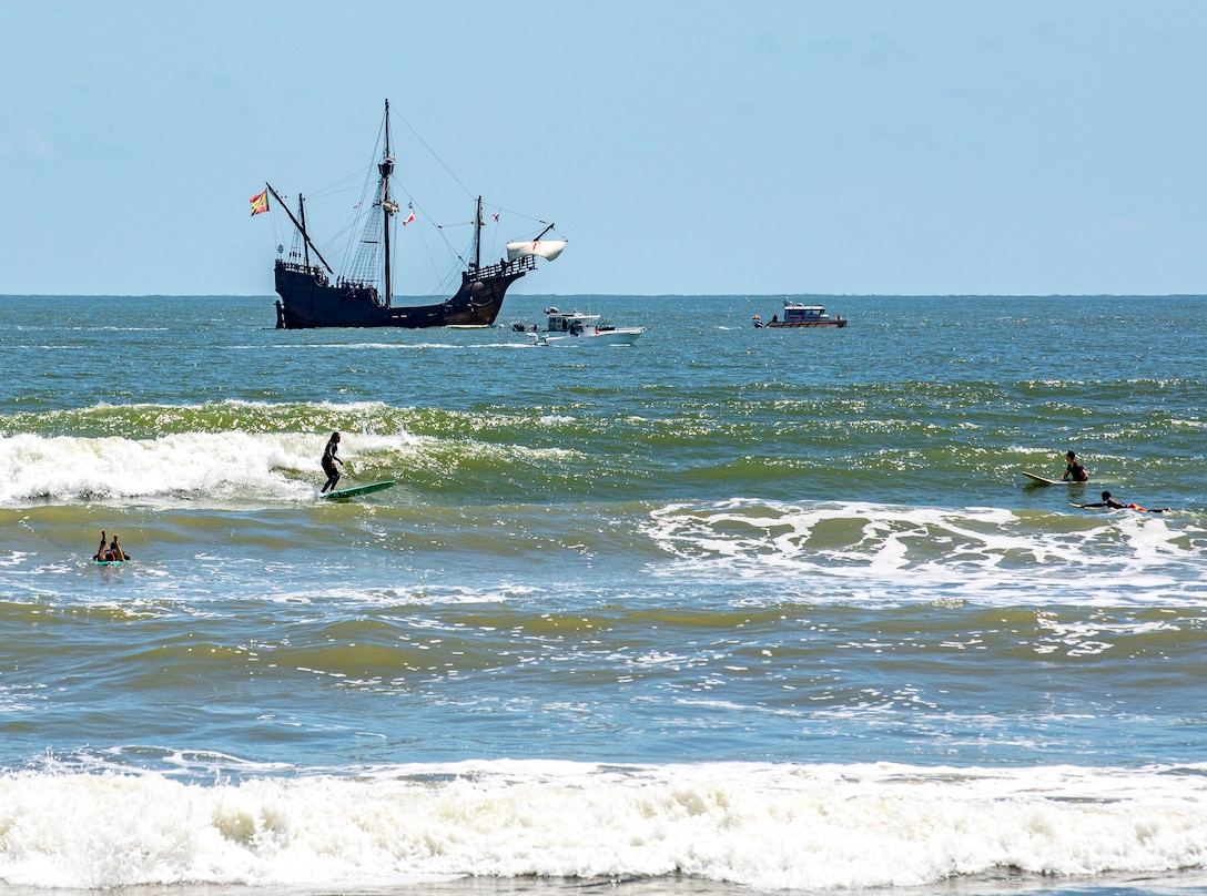 Surfers ride waves as Nao Santa Maria, replica of the Nao Santa María of Christopher Columbus, transits the Gulf of Mexico off the coast of Galveston Beach during Tall Ships Challenge Galveston 2023.