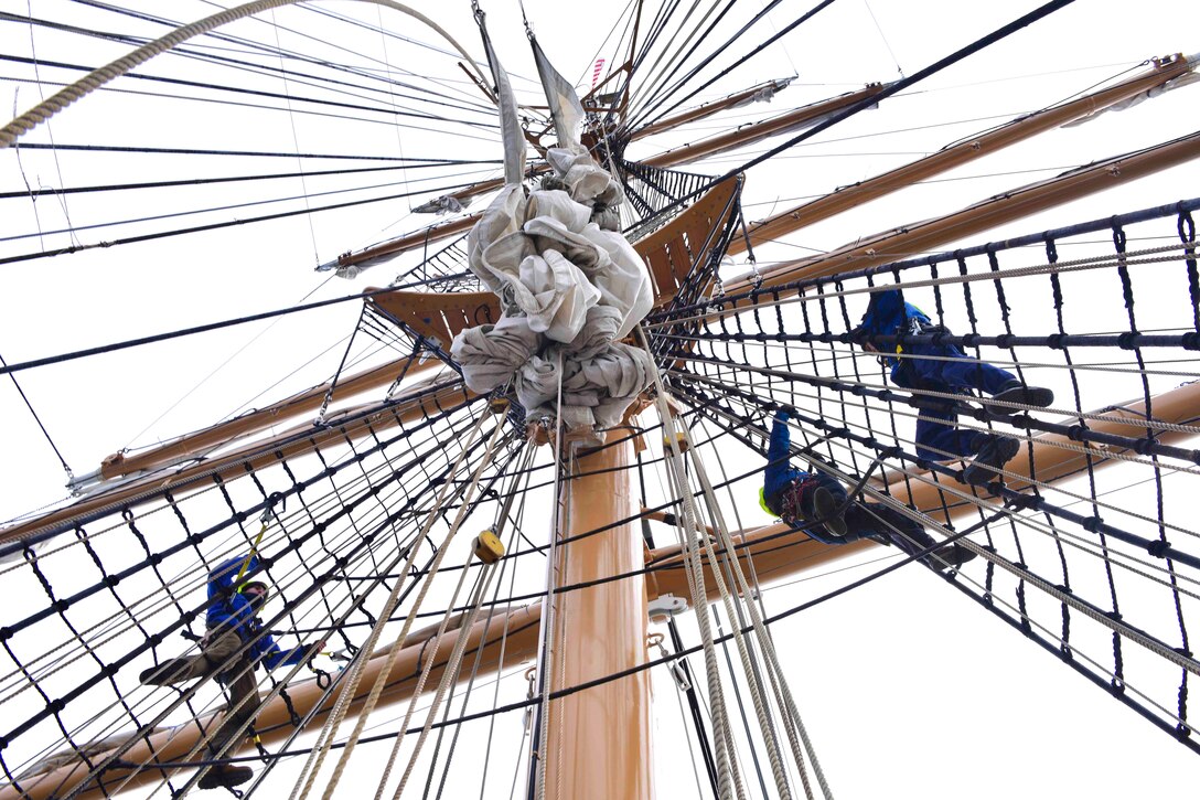 Coast Guard personnel seen from below climb the rigging of a tall ship.