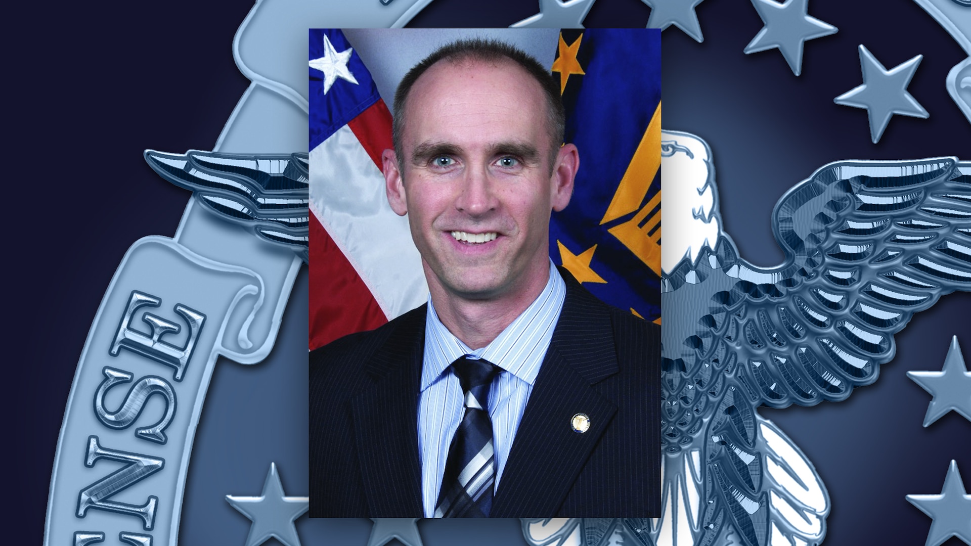 A professional headshot of a man sitting in front of the American flag and the Defense Logistics Agency flag.