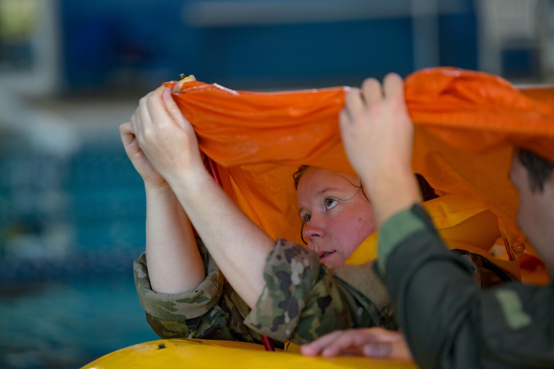 Two airmen hold a piece of orange material above their heads while floating on a yellow device in a pool.