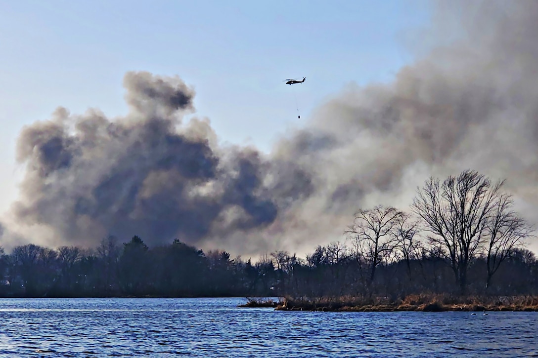 An Army helicopter with a bucket attached by a line flies above gray smoke in a forested area bordering water.