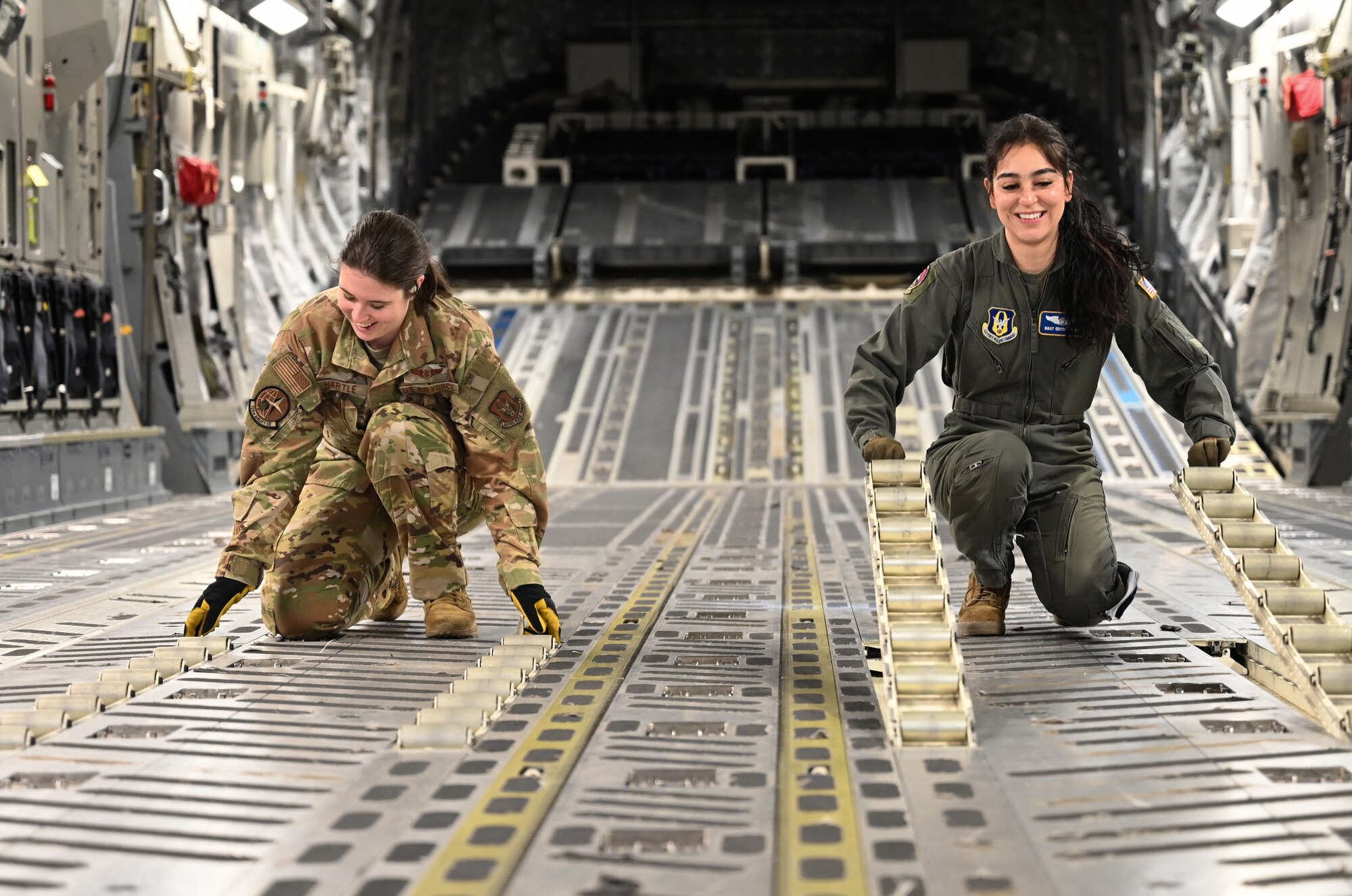 Two female loadmasters in flight suits flip rollers on the floor of the inside of a C-17 Globemaster.