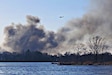 An Army helicopter with a bucket attached by a line flies above gray smoke in a forested area bordering water.