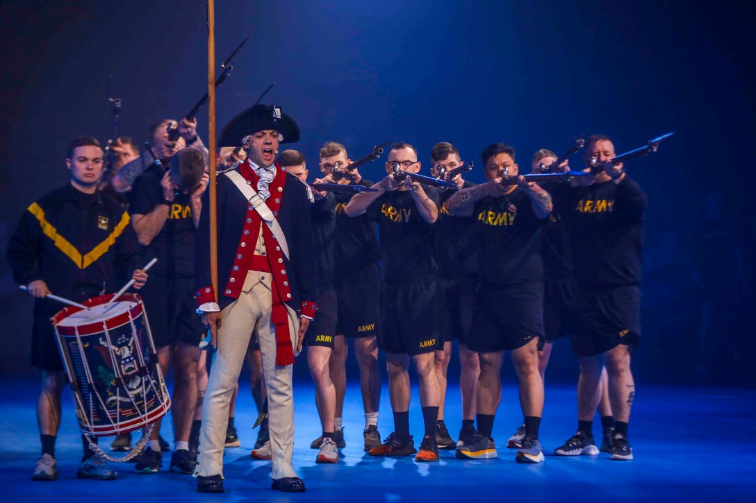 A soldier sings on stage while standing in front of group of other soldiers pointing rifles and one playing a drum.