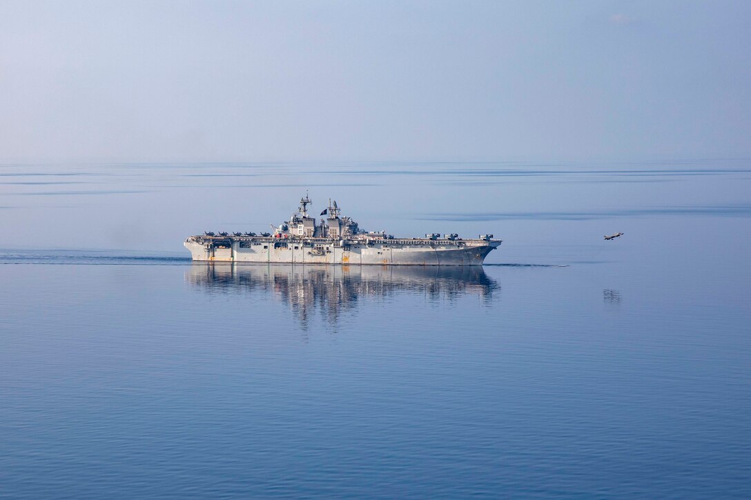 A Marine Corps aircraft flies in front of ship traveling in blue water.