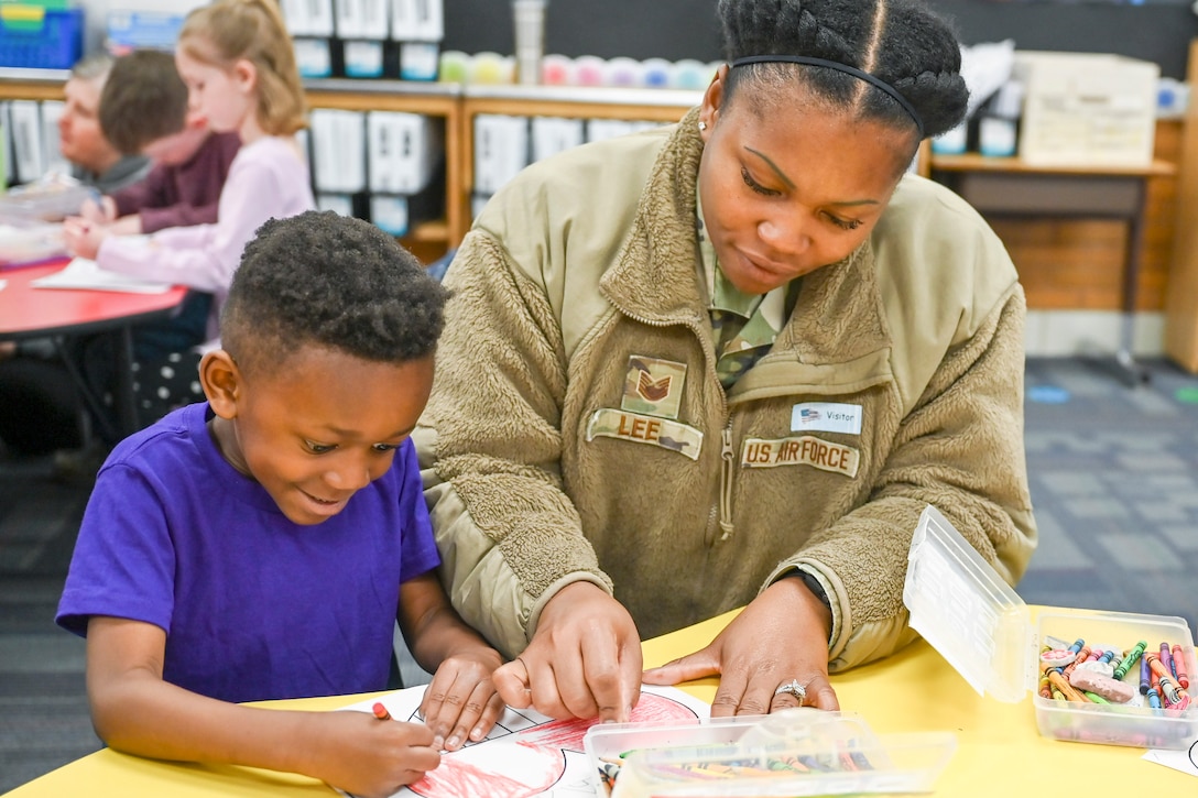 An airman and son color while sitting next to each other in a classroom with fellow students and parents in the background.