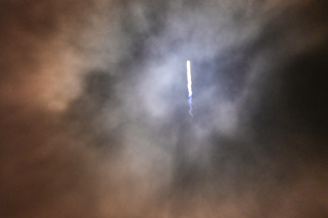 A rocket passes through clouds in a dark sky as seen in the distance.