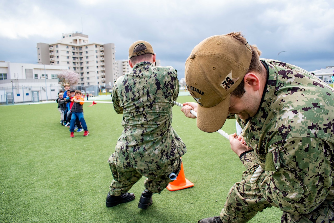 Sailors play tug-of-war with students on a field.
