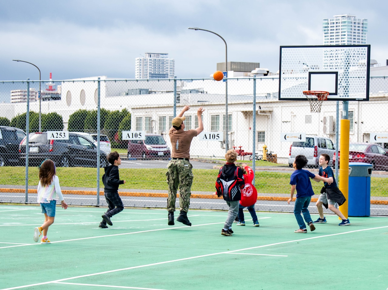 A sailor shoots a ball while playing basketball with elementary students.