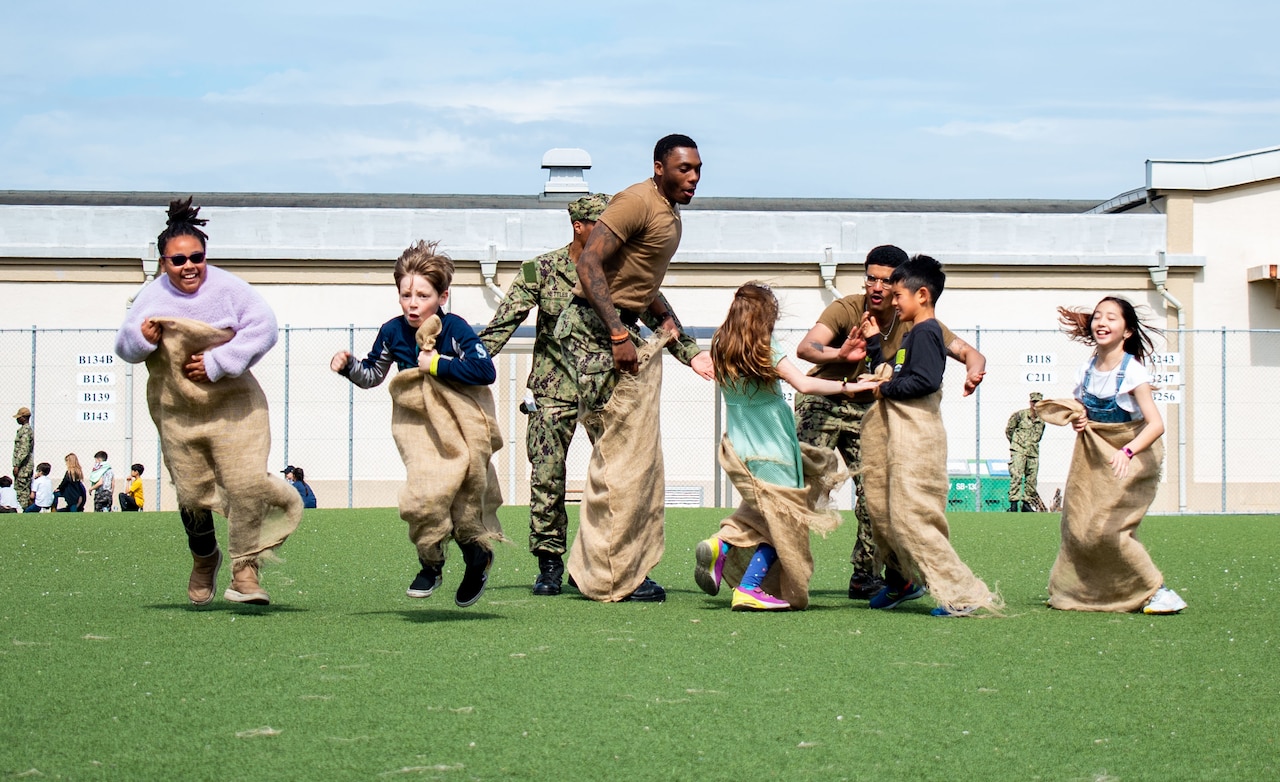 A sailor jumps in potato sack with elementary students in a field.