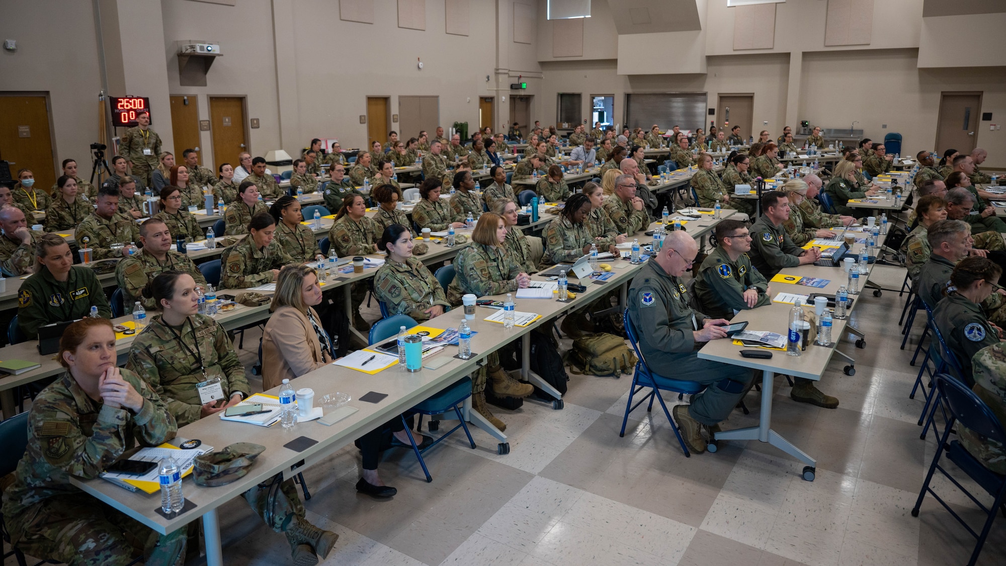 A large group of Airmen sit in a room at tables arranged horizontally across the frame