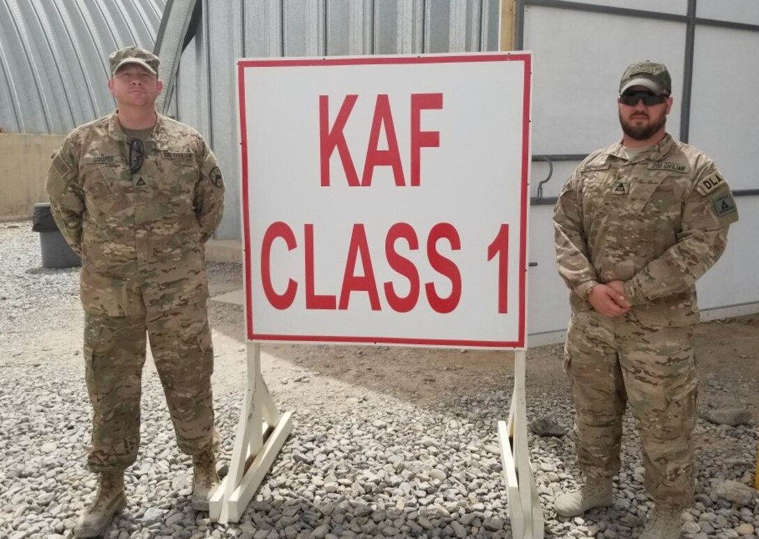 Two men on dark skinned, one light skinned in military OCP camouflaged uniforms stand in front of a bright red and white Class 1 sign in a desert environment.