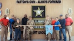 Two people in military OCP camouflaged uniforms stand with nine civilians in business casual attire holding a Guide-on DLA unit flag in front of a Army building.