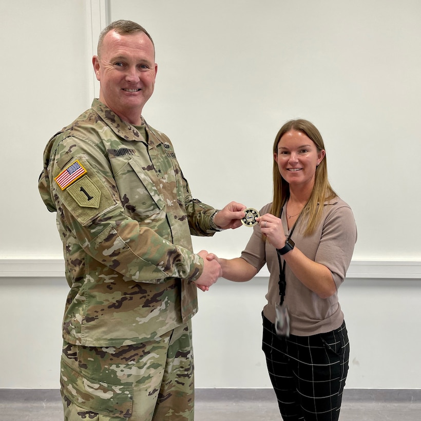 A tall man in military OCP camouflaged uniform with a buzz cut hands a coin to a short woman with brown hair, wearing a tan shirt and black pants.