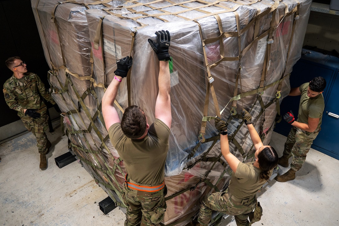 Four airmen strap down a large pallet of boxes covered in plastic.