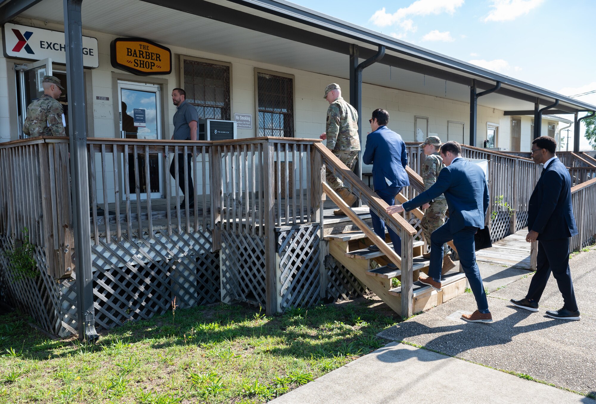 A military legislative assistant, military affairs director, and congressman follow an Air Force military commander up some wooden stairs outside leading to a shoppette