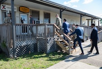 A military legislative assistant, military affairs director, and congressman follow an Air Force military commander up some wooden stairs outside leading to a shoppette