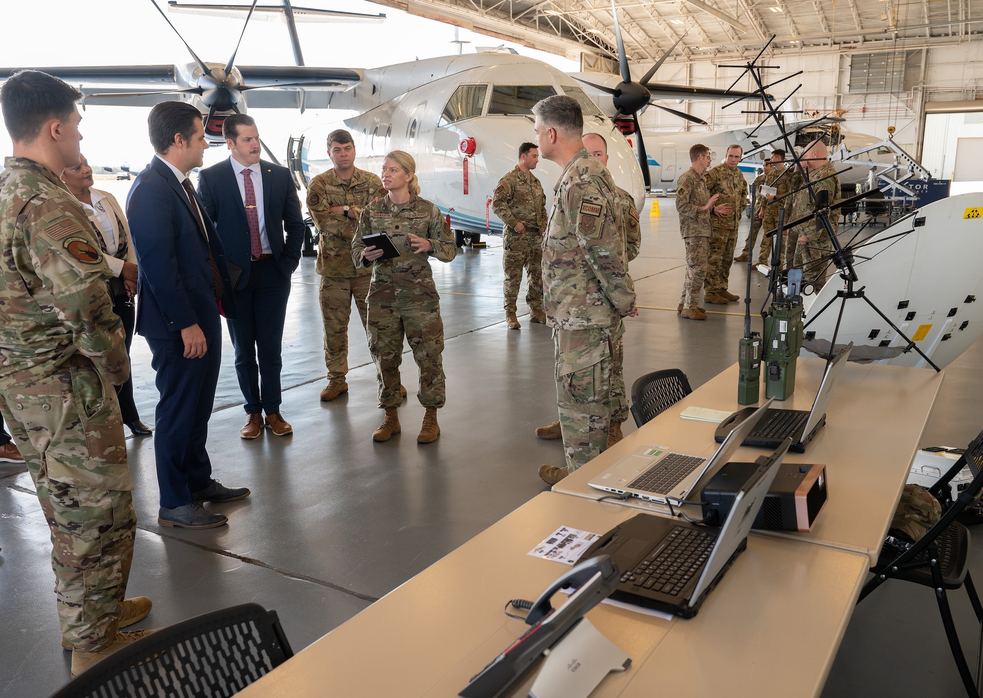 In the foreground is tactical communications equipment displayed on a table and behind it are senior leaders talking in a C-146A Wolfhound hangar
