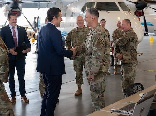 Congressman and military member shake hands in a hangar. Other senior leadership stand around with C-146A aircraft behind them.