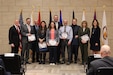 Ten people in business attire stand in front of flags and hold certificates of appreciation and award for service.