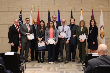 Ten people in business attire stand in front of flags and hold certificates of appreciation and award for service.