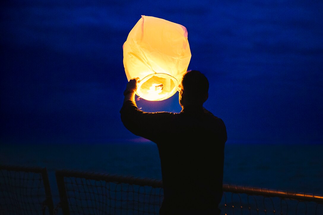 A sailor holds a lighted paper lantern over a ship's deck against a dark blue sky.