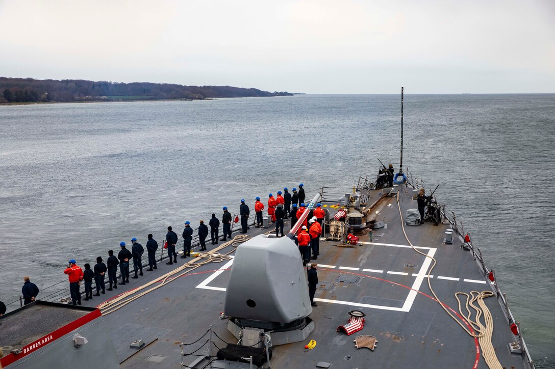 Sailors stand on deck as a ship sails into open sea.