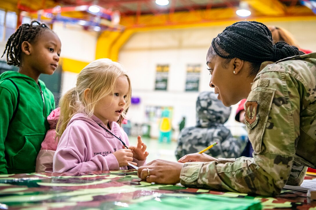 An airman sitting at a table speaks to two children.