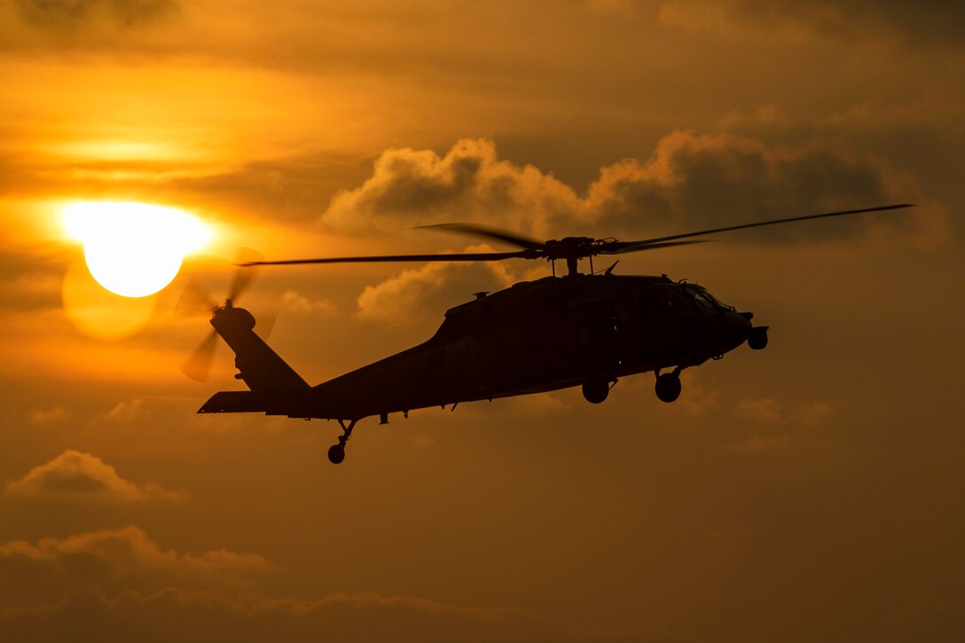 A helicopter flies near clouds as the sun sets to its left as shown in silhouette.