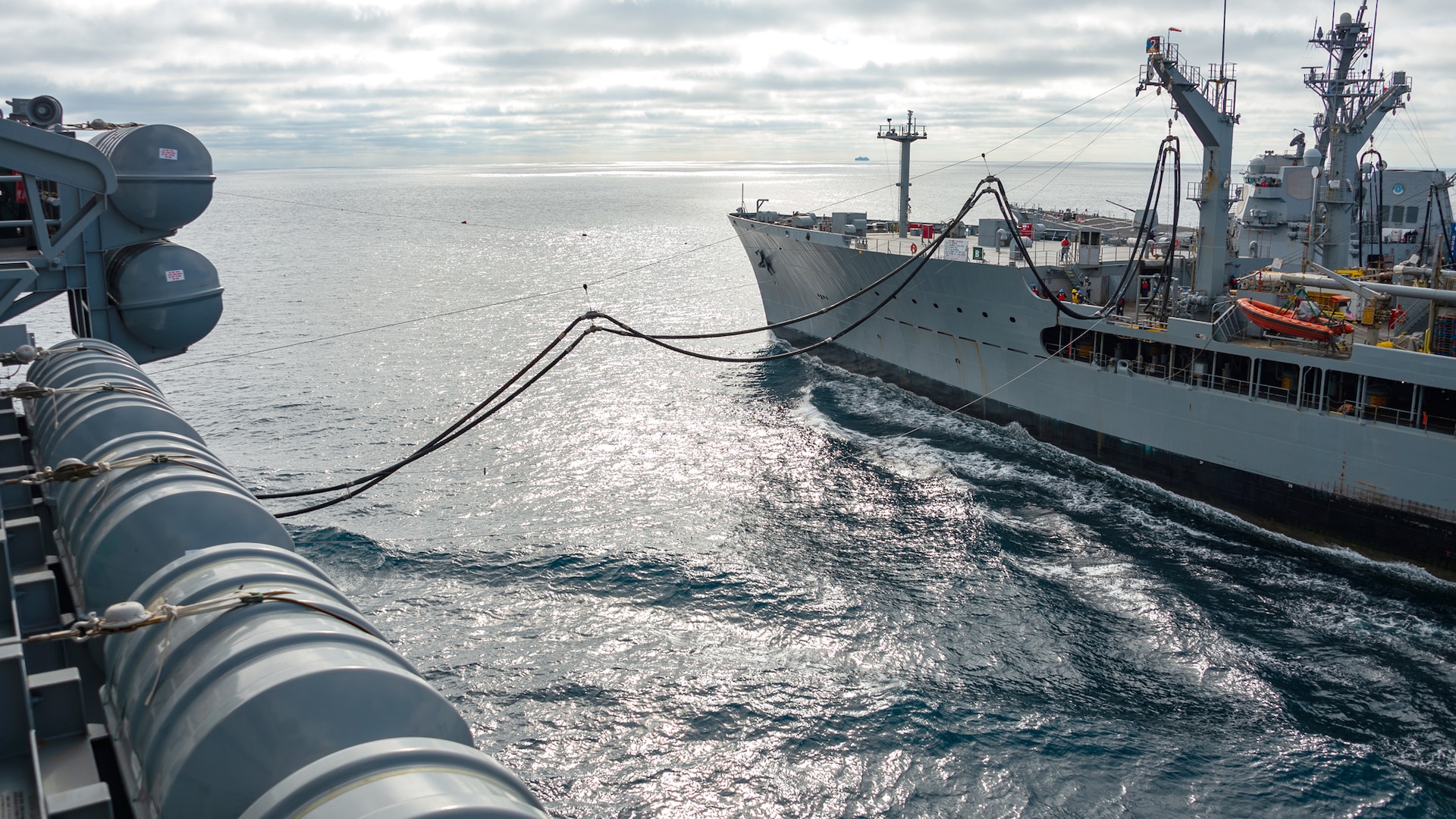 two Navy ships at sea with a fuel hose strung between them